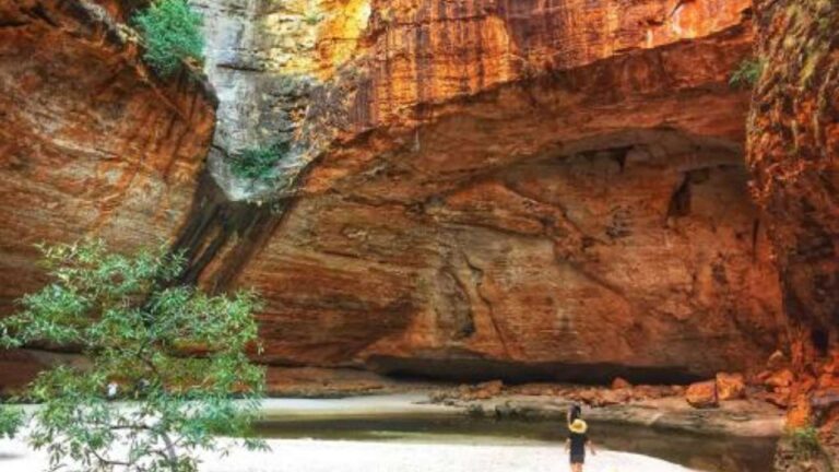 Person standing in big canyon of red rock, with sandy beach and waterhole at bottom